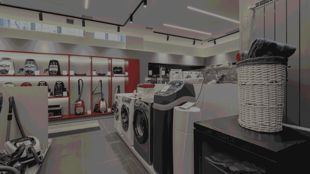 Interior of an appliance store displaying washing machines, vacuum cleaners, and other electronics.