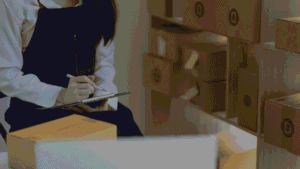 Woman in denim apron writing on clipboard beside shelves of packages