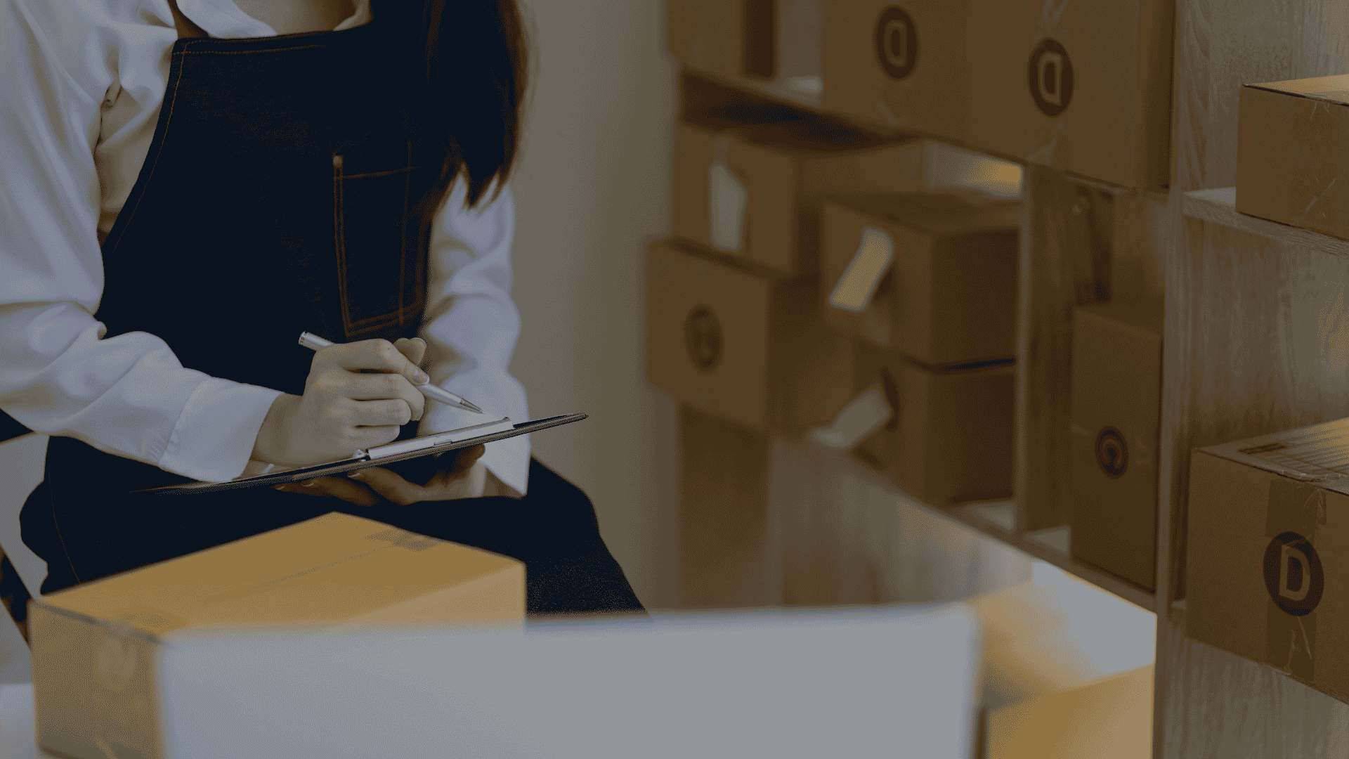 Woman in denim apron writing on clipboard beside shelves of packages