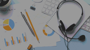 Flat-lay of a desk with a customer support headset, keyboard, printed charts, and stationery on a light blue background.