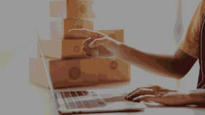 Person in a yellow shirt points toward a stack of cardboard shipping boxes while using a laptop.