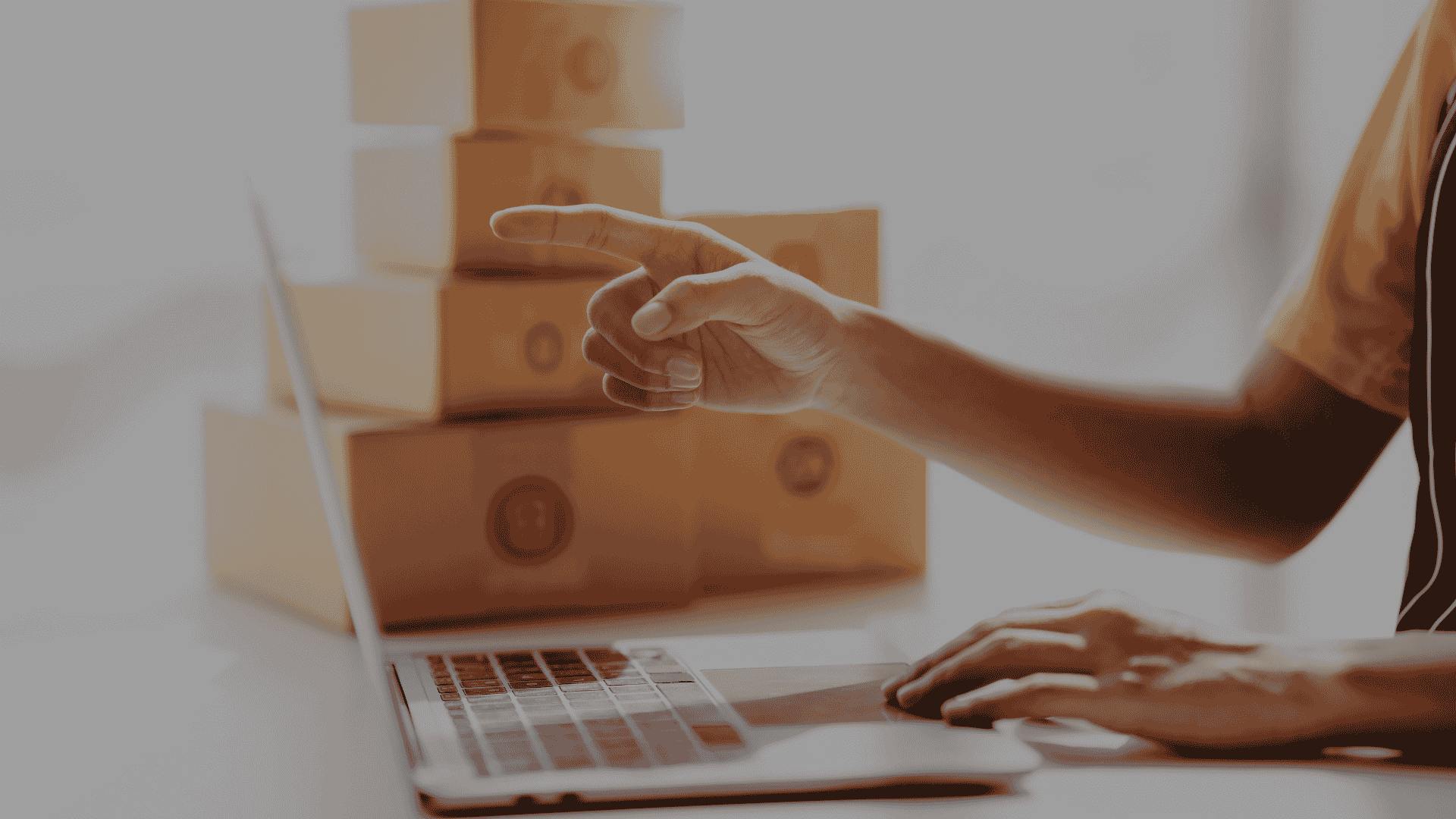 Person in a yellow shirt points toward a stack of cardboard shipping boxes while using a laptop.
